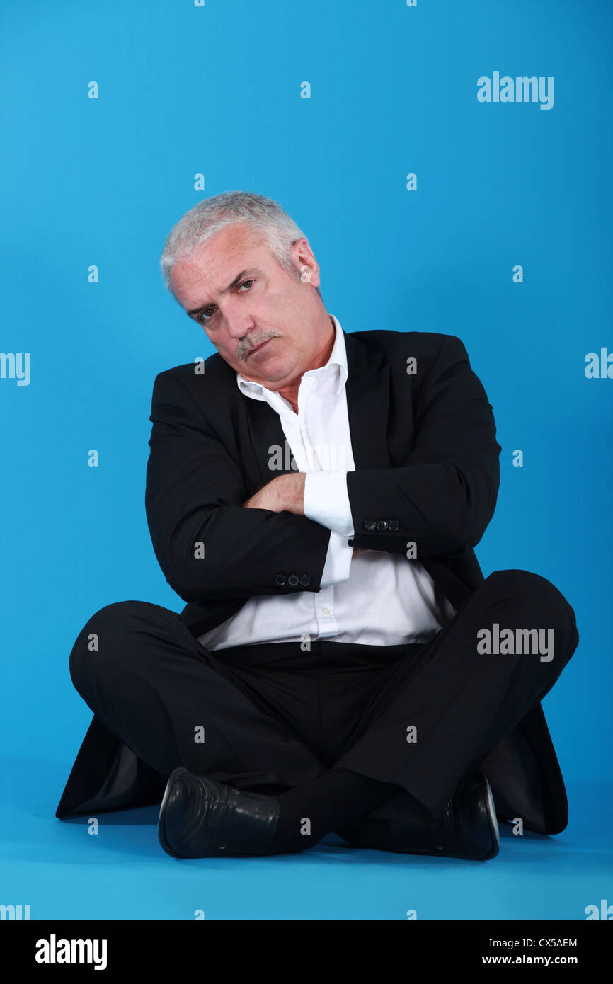 middle-aged man sitting cross-legged and cross-armed against blue background Stock Photo