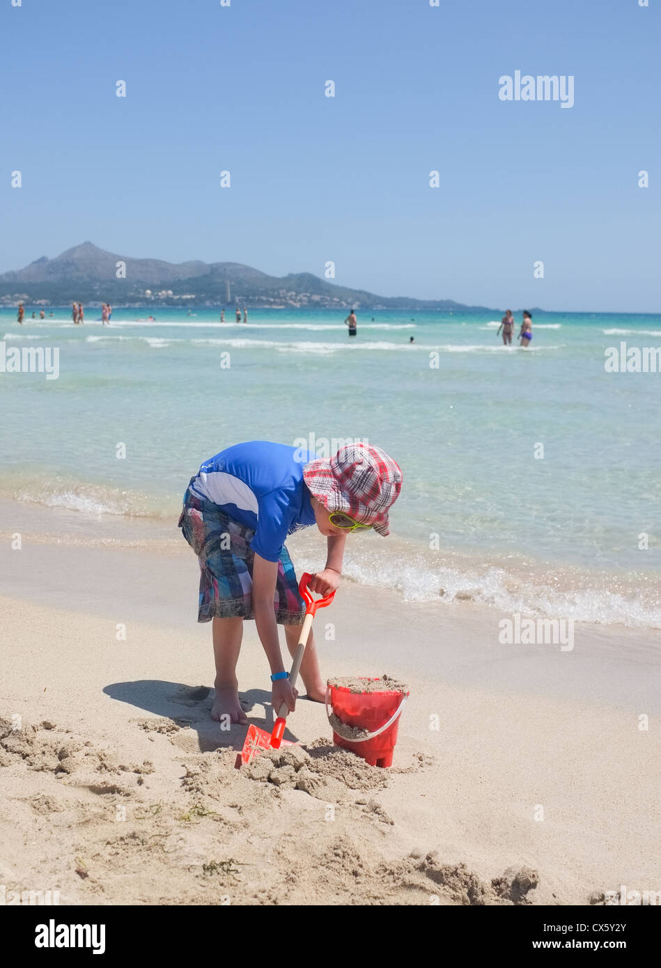 A young boy digs in the sand at alcudia beach Majorca wearing sun protection hat and vest Stock Photo