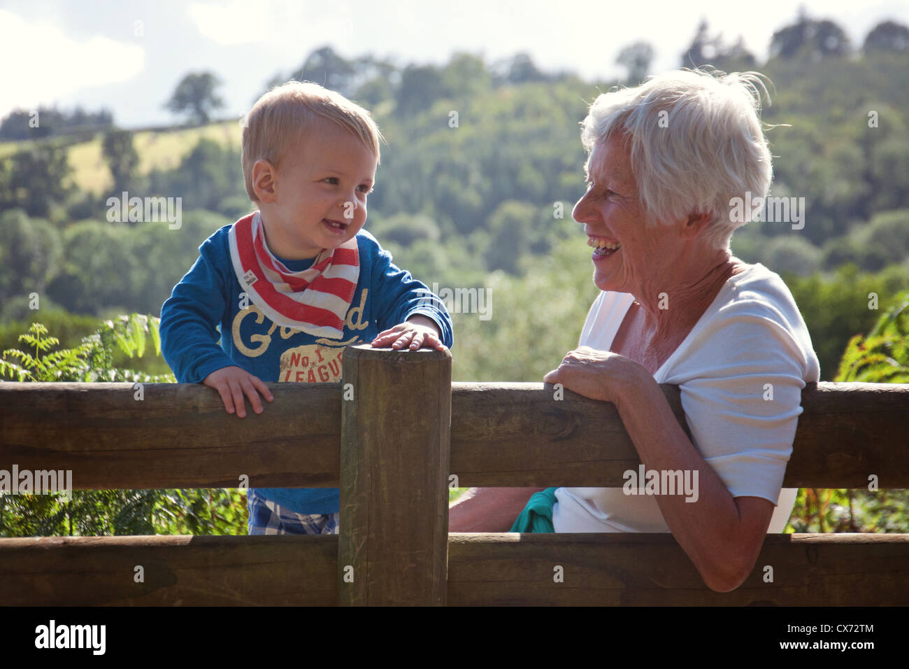 Elderly woman resting her arm over the back of a bench while a toddler boy stands next to her on the bench in a country setting Stock Photo