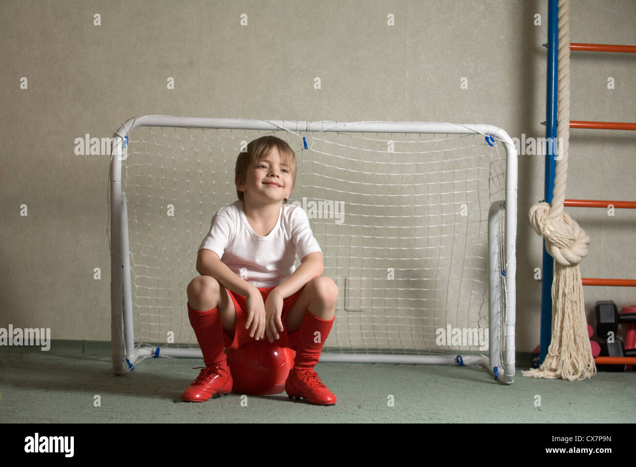 A young boy sitting on a soccer ball in front of a soccer goal Stock Photo
