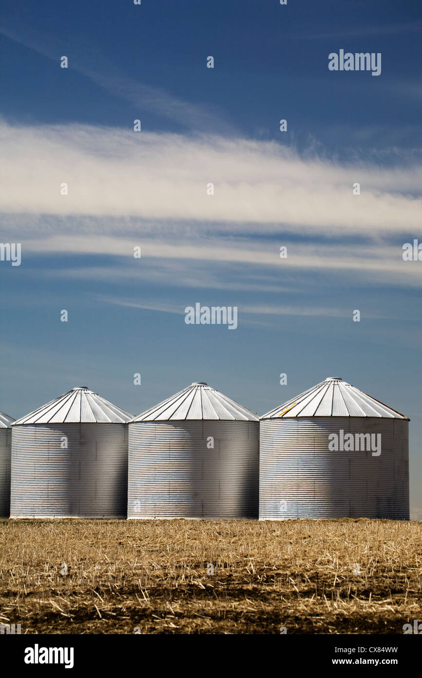 Three Shiny Metal Grain Bins In Stubble Field With Blue Sky And Clouds