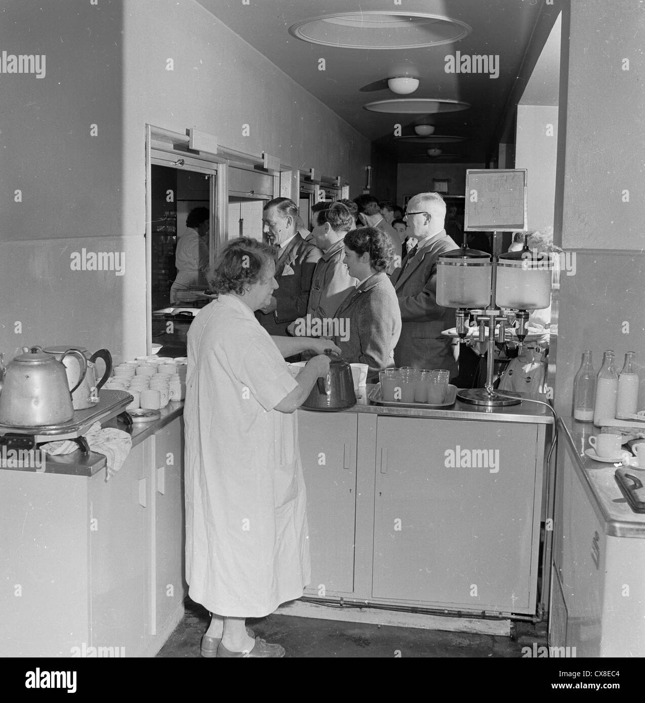 England,1950s. Factory workers wait at the canteen hatch for food, while a female dinner lady pours tea. Stock Photo