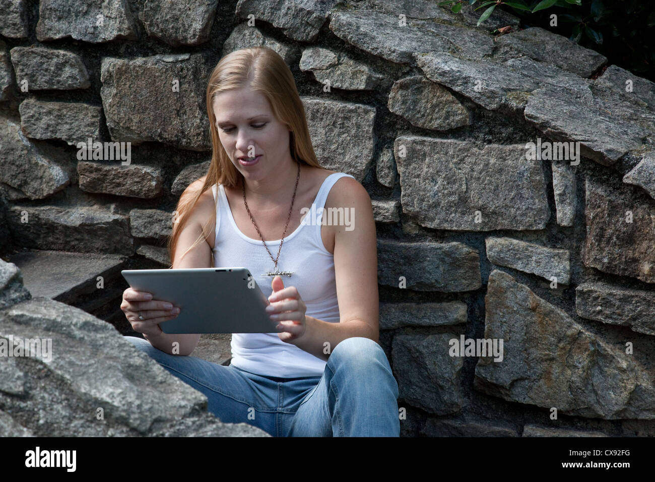 Pretty blond woman sitting outside on stone steps and working on a computer tablet in the warm sunshine. Stock Photo