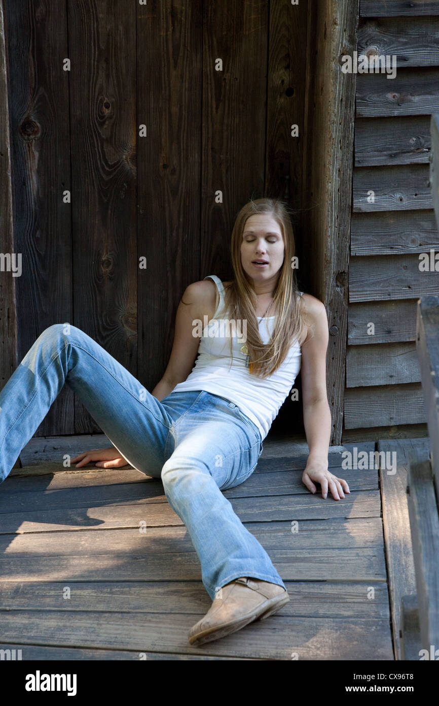 Pretty blond woman outside on a wooden walkway with an old weathered door behind her, looking like she has passed out Stock Photo