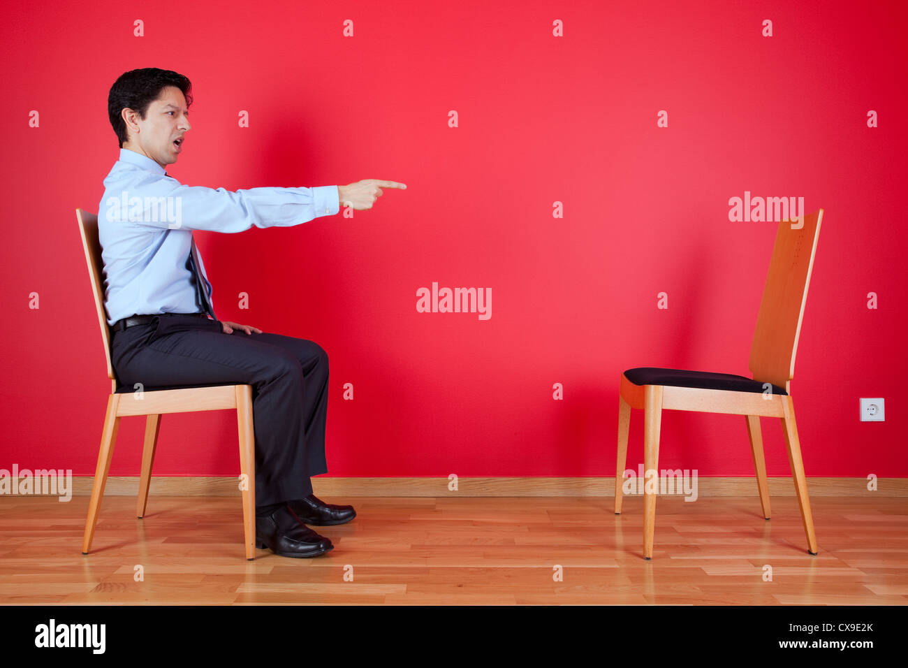 Businessman sited next to a red wall pointing to a empty chair Stock Photo