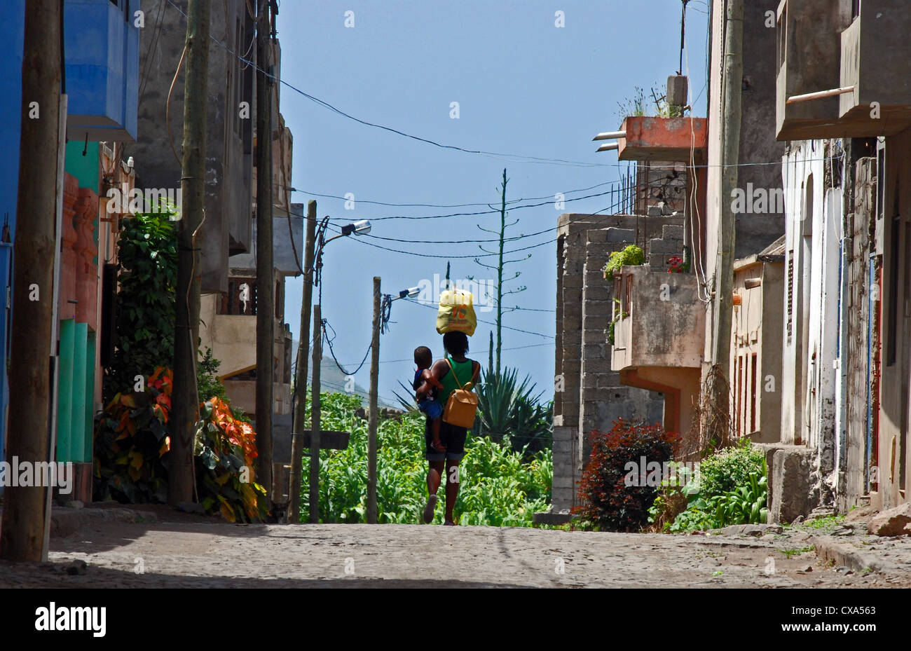 A Cape Verdean girl walks along a cobbled street carrying a young child on her hip and a shopping bag on her head. Stock Photo