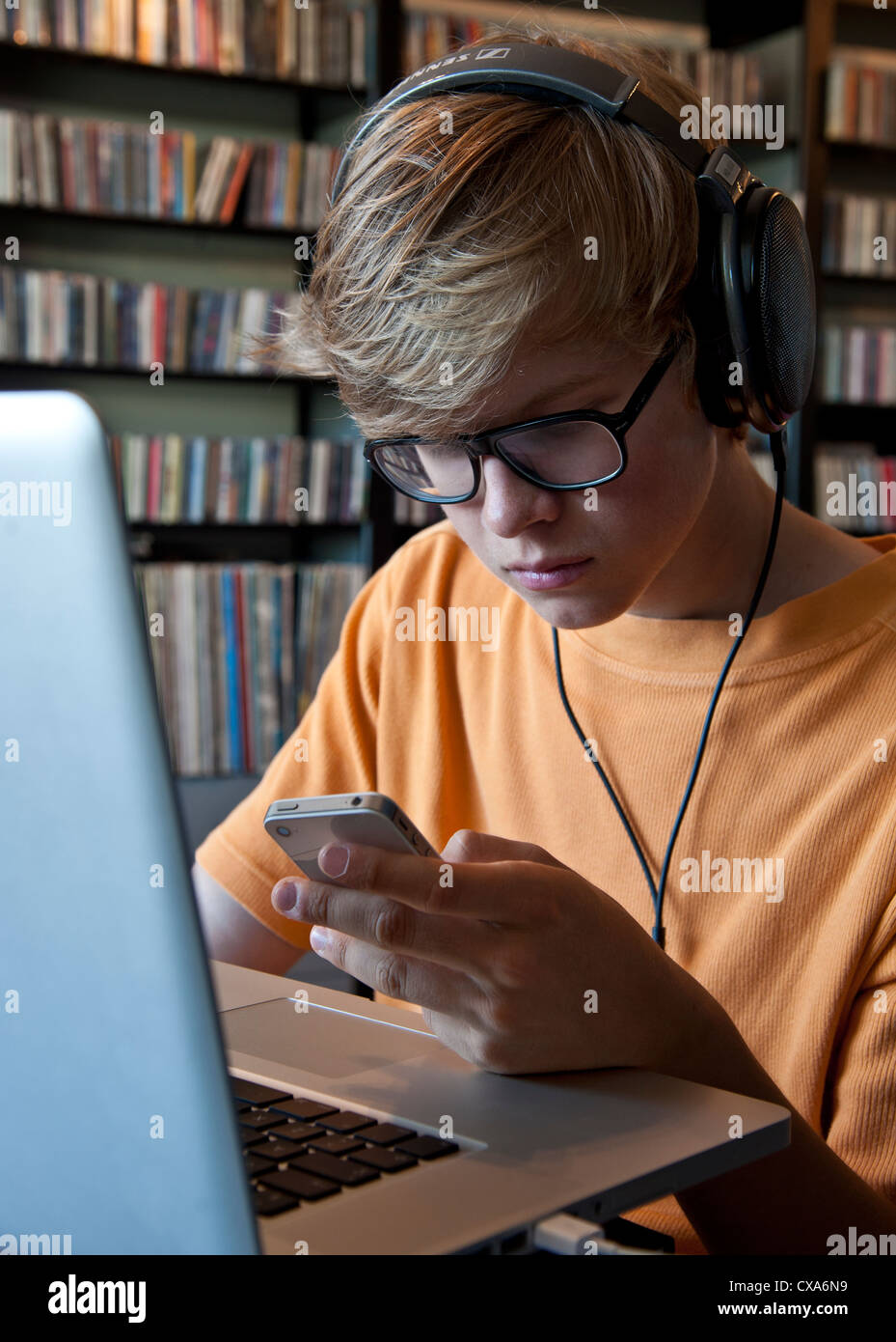 Studious relaxed teenage school boy wearing headphones using his Apple iPhone smartphone and laptop computer Stock Photo