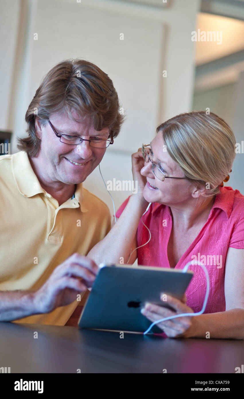 Attractive mature couple at home listening to their iPad tablet computer together, in light contemporary living room Stock Photo