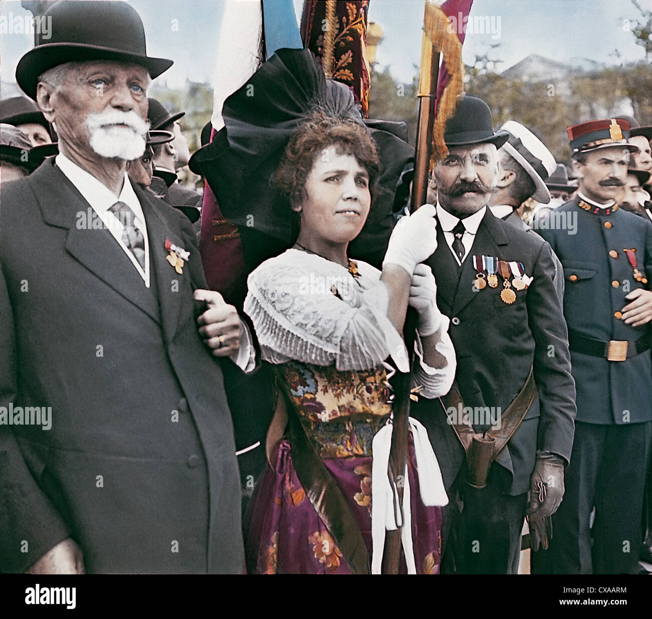 Colorized photo of a group of people at the Armistice Parade on Armistice Day, Paris, France, November 11th, 1918. A soldier and two plain-clothed men, decorated with medals, stand on either side of a woman holding a flag. (Photo by Burton Holmes) Stock Photo