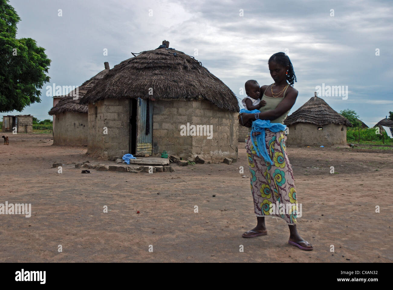 A young senegalese mother holds her baby, stands outside her breeze block and thatch construction hut. Stock Photo