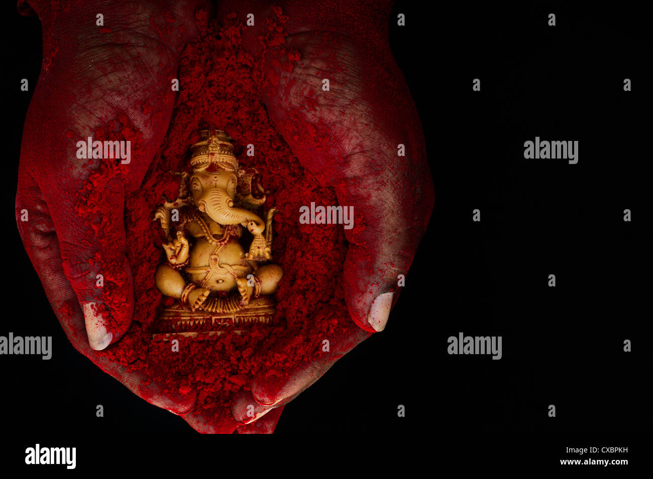 Hindu Elephant God. Red powder covered indian mans hand holding Lord Ganesha statue against black background Stock Photo