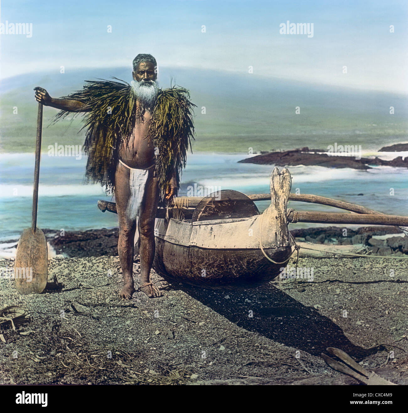 Colorized portrait of a native Hawaiian man standing next to an outrigger  canoe on a beach, Hawaii, 1898. The man wears a ti leaf rain cape and holds  a paddle. (Photo by