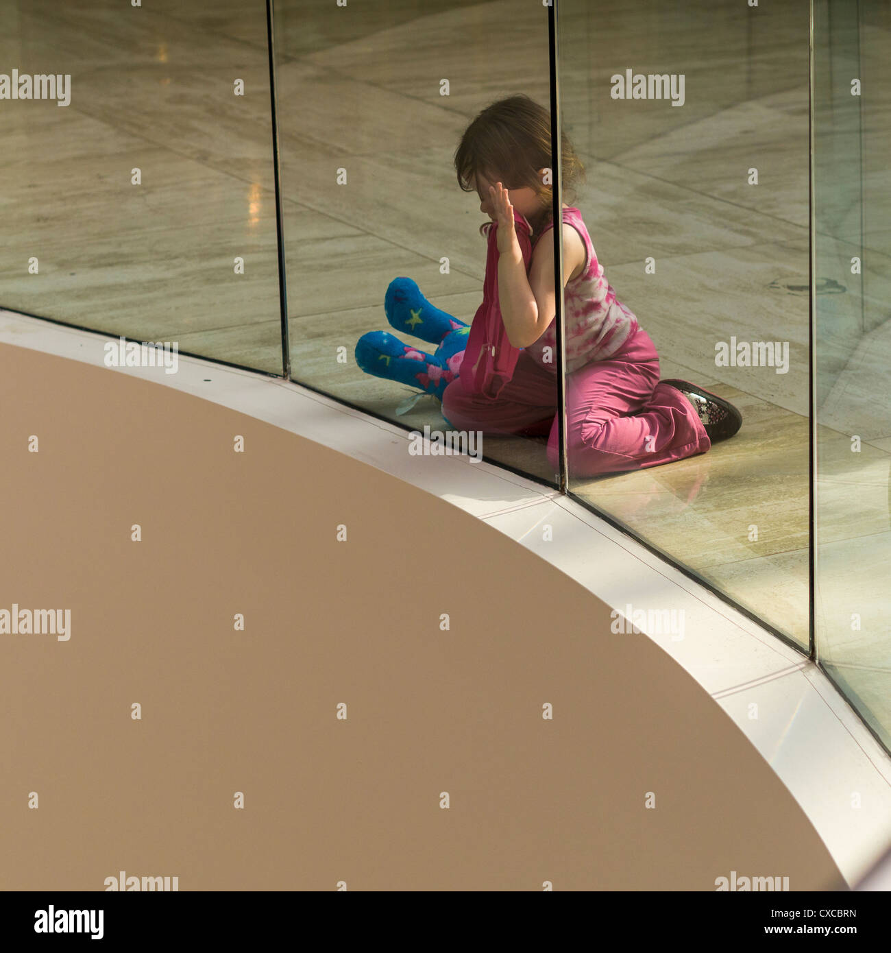 Watching. A young girl with a stuffed animal sits on the mall floor and watches something below while pressed up against a glass Stock Photo