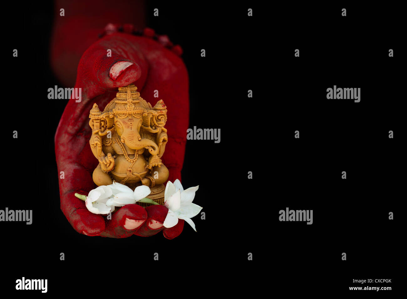 Hindu Elephant God. Red powder covered indian mans hand holding Lord Ganesha statue and jasmine flowers against black background Stock Photo