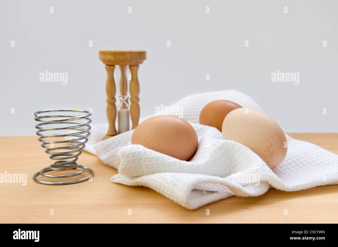 Still life image of egg timer, egg sand egg cup on napkin on top of wooden chopping board against a white background Stock Photo