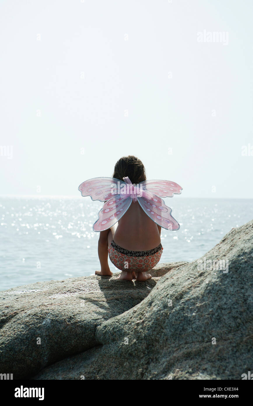 Girl wearing butterfly costume, crouching on rock and looking at ocean Stock Photo