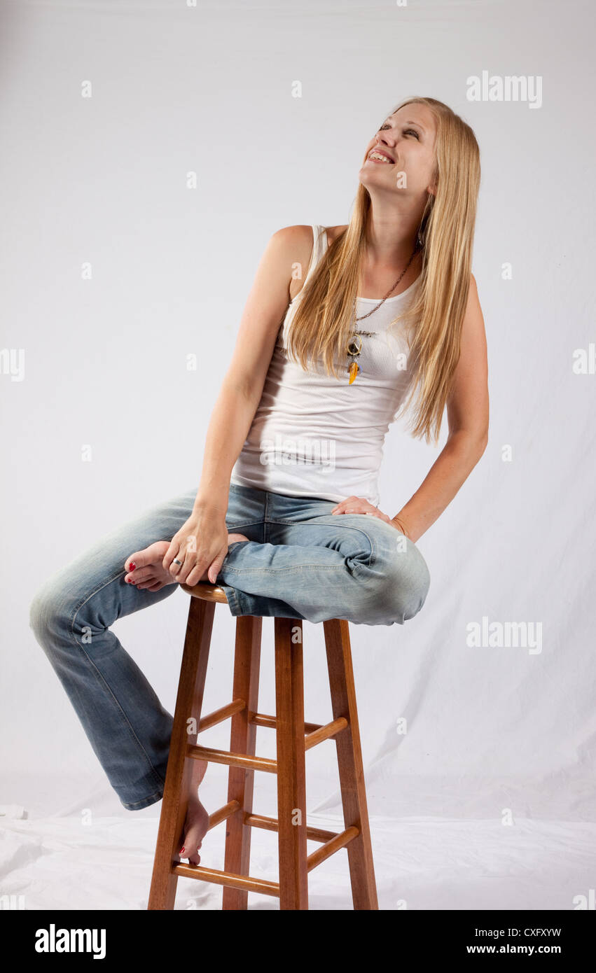 Pretty blond woman sitting on a wooden stool with one leg across the stool, and looking up with a happy, glad smile Stock Photo