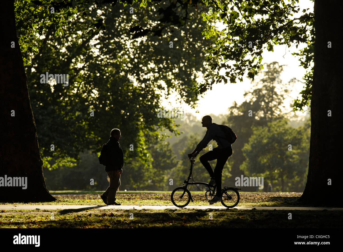 A man walks though Hyde Park whilst another cycles on a foldup bike in