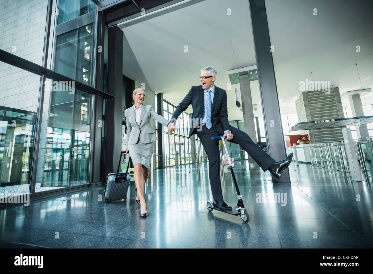 Germany, Stuttgart, Businesswoman with wheeled luggage, man riding scooter at office building Stock Photo