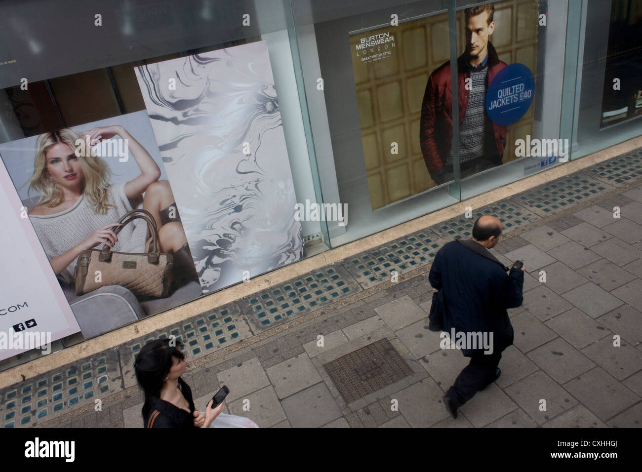 Two smartphone consumers walk along a London street next to fashion ad posters. Stock Photo