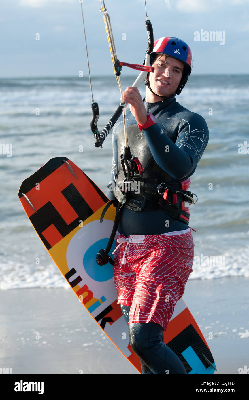 Smiling, wet, young man in wetsuit, with helmet and kiteboard holds up the bar of a kitesurf kite Stock Photo