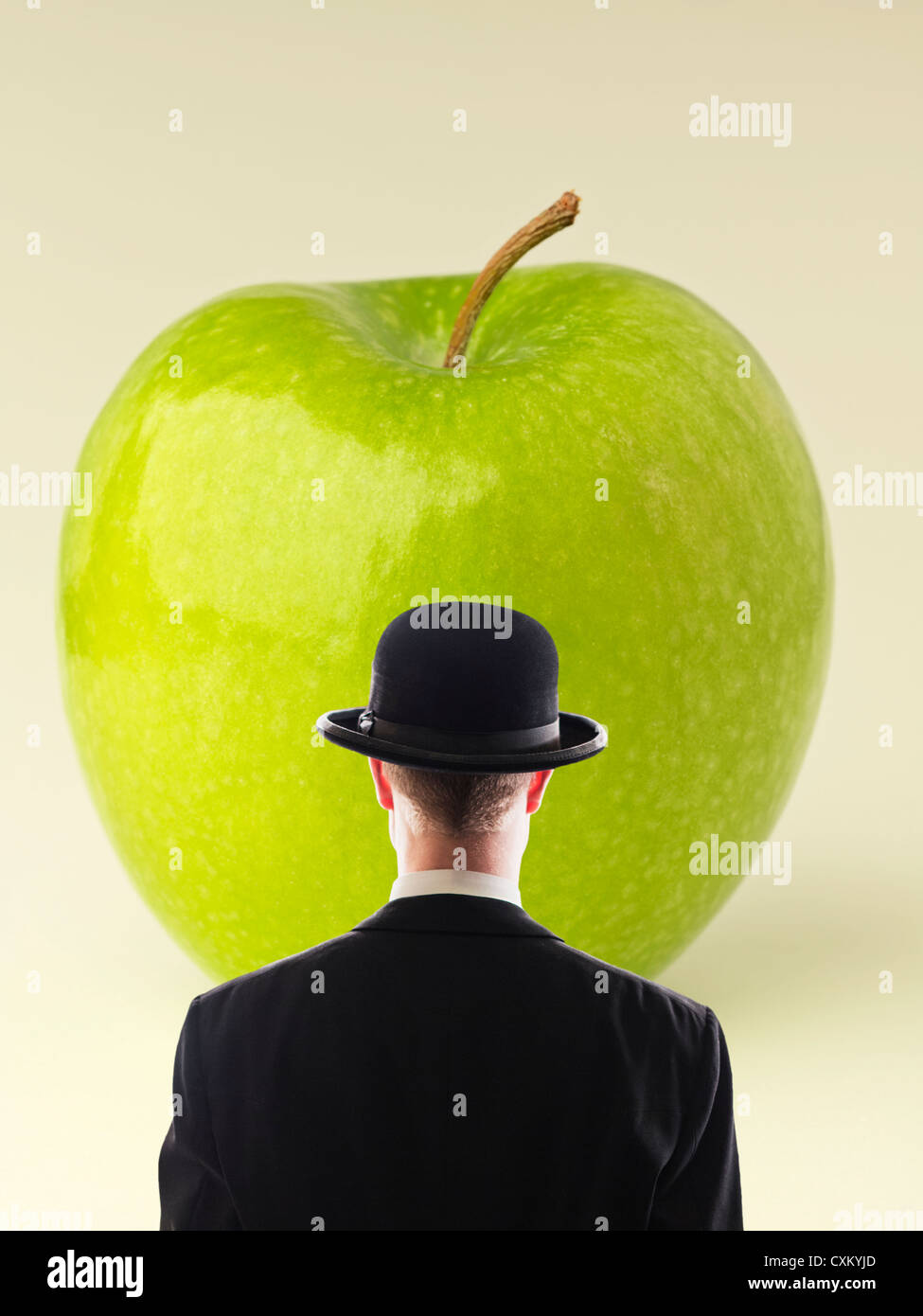 Man in black suit and bowler hat facing a large green apple. Based on the surreal painter Rene Magritte. Stock Photo