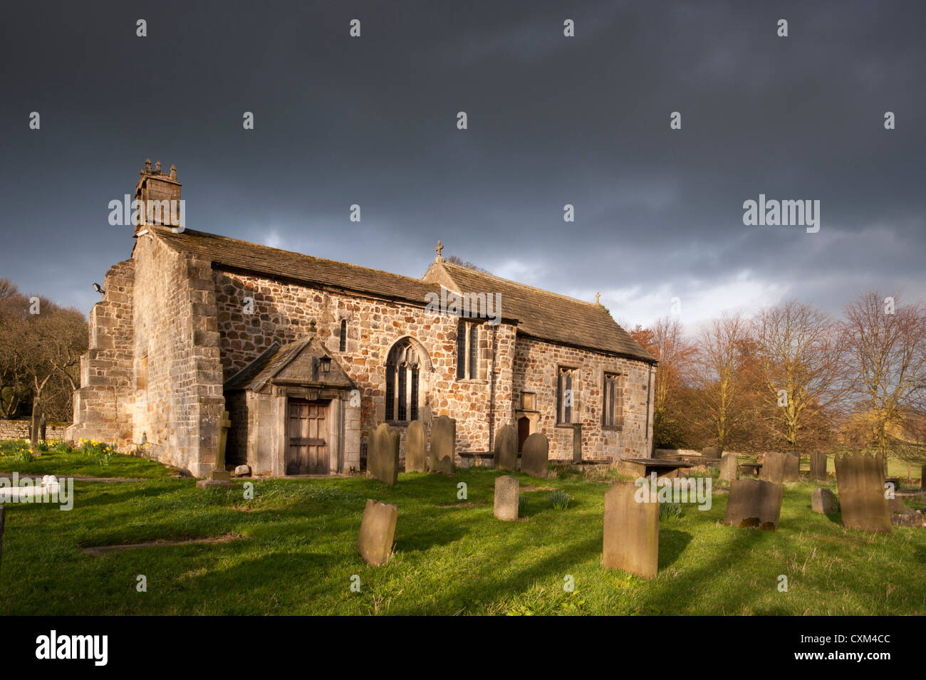 Exterior of historic sunlit All Saints Church & churchyard headstones in quiet scenic countryside under dark sky, Weston, North Yorkshire, England, UK Stock Photo