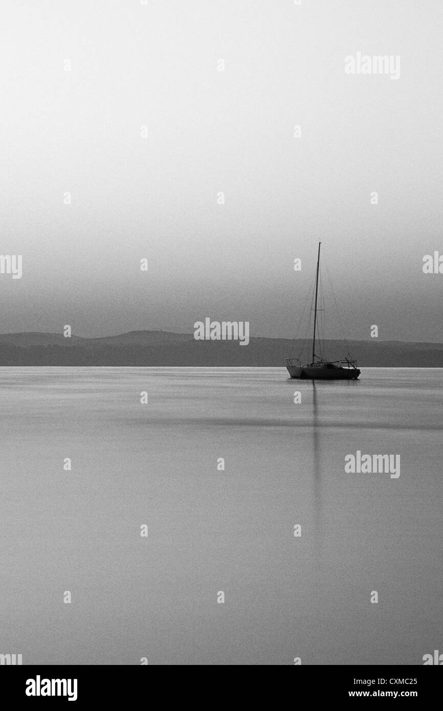View of a Sailing Boat moored on the water at dawn. The boat's mast is reflected in the water Stock Photo