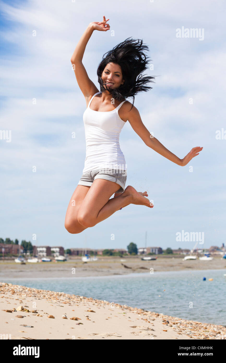 Joyful, happy, excited young woman with long dark hair jumping for joy on beach in summer Stock Photo