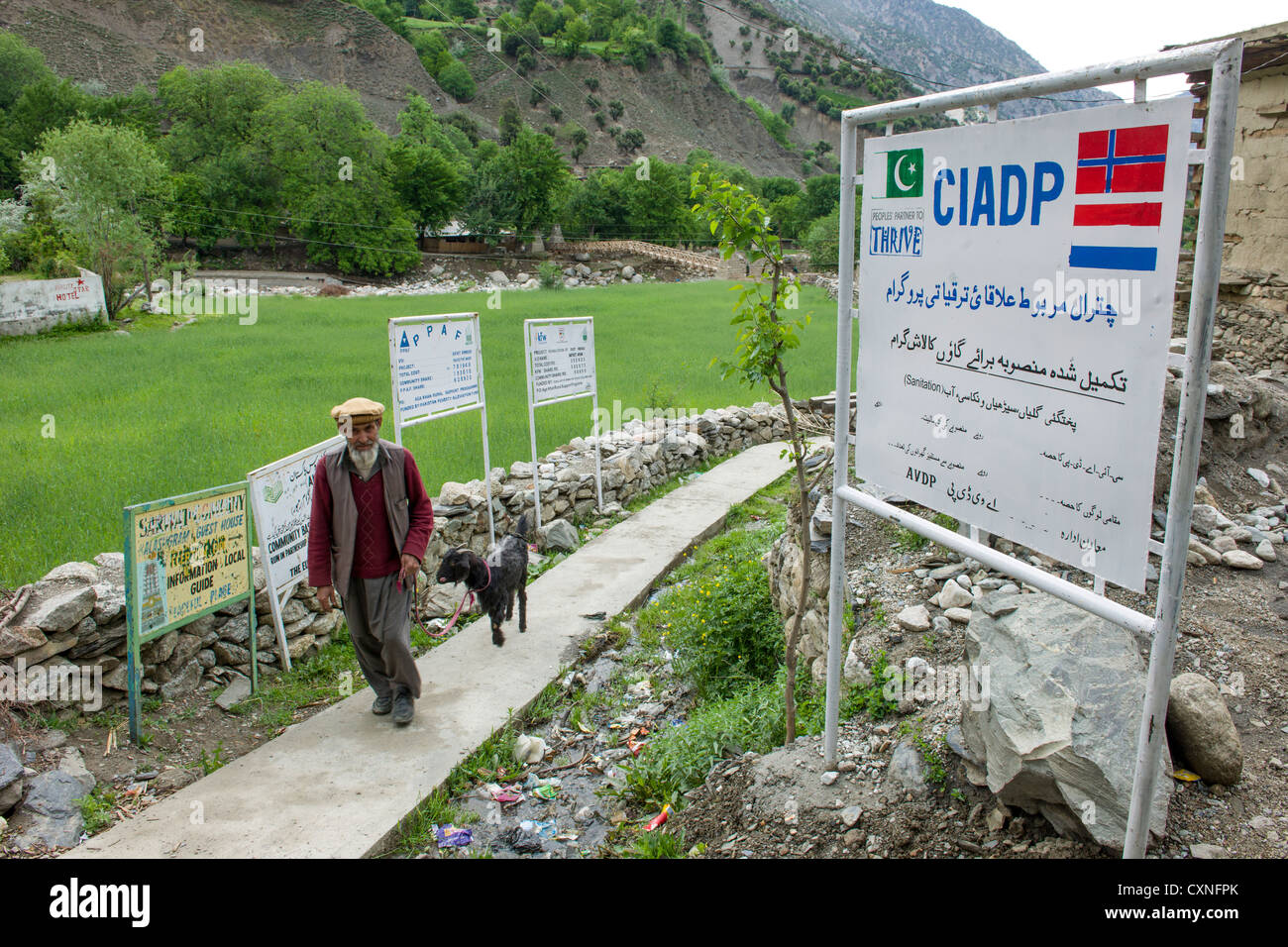 Muslim man leading his goat past signs promoting the projects of various development agencies, Rumbur Valley, Chitral, Khyber-Pakhtunkhwa, Pakistan Stock Photo