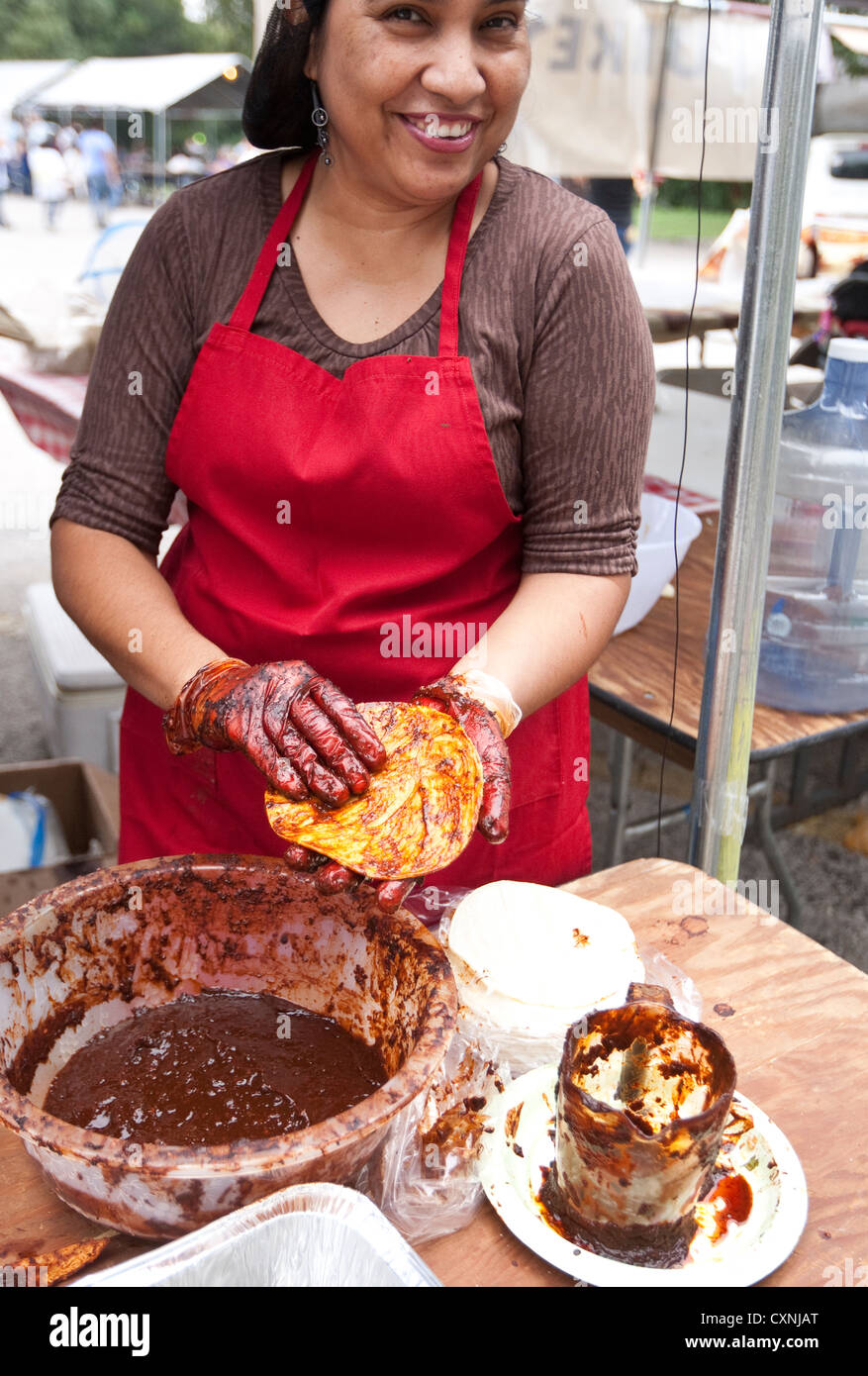 Hispanic woman prepares traditional Mexican enchiladas by covering corn tortilla with red chile sauce at outdoor church festival Stock Photo
