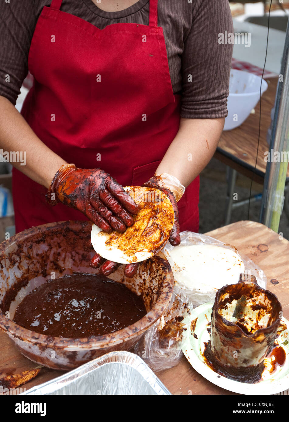 Hispanic woman prepares traditional Mexican enchiladas by covering corn tortilla with red chile sauce at outdoor church festival Stock Photo