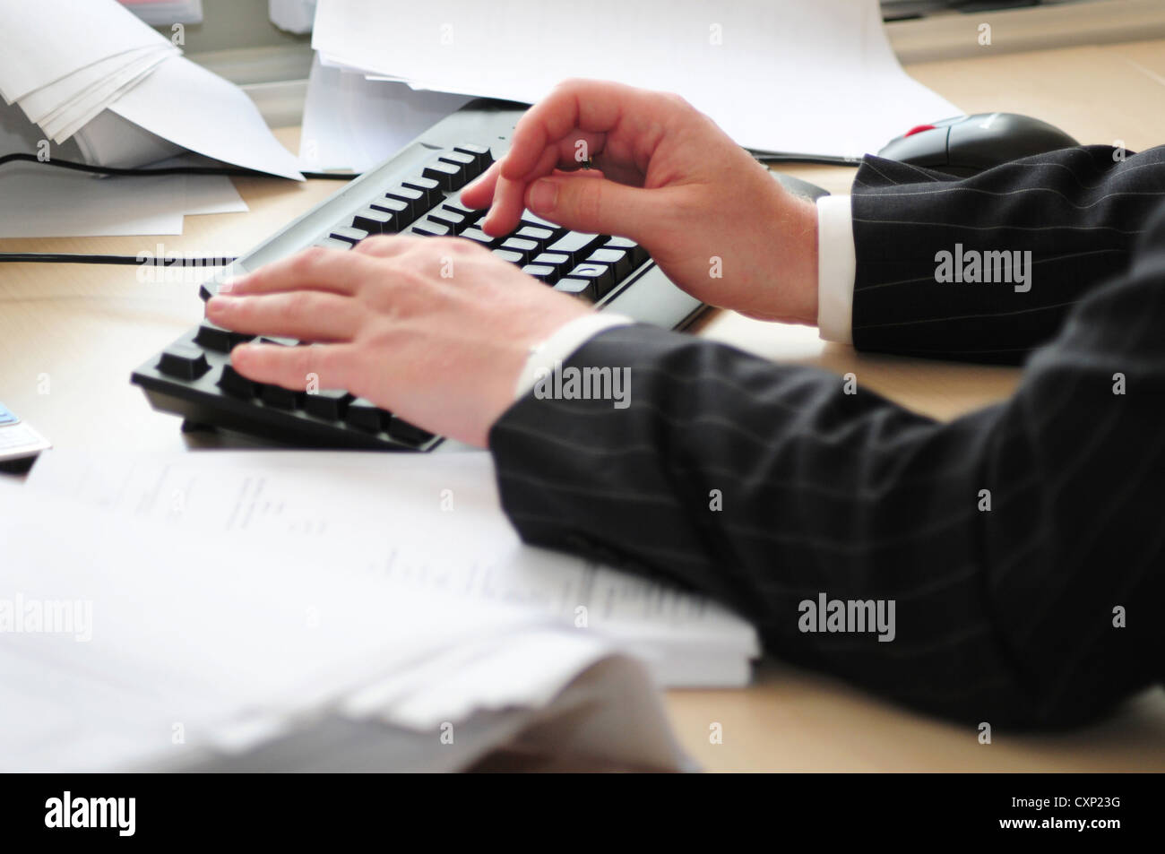 Computer keyboard on a busy office desk with selectivly focused male hands in pinstripe suit typing surrounded by papers. Stock Photo