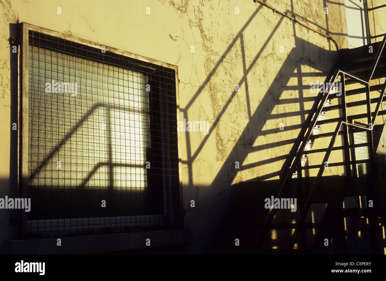 Detail of metal fire escape stairs casting shadows on pale wall with security window in evening light Stock Photo