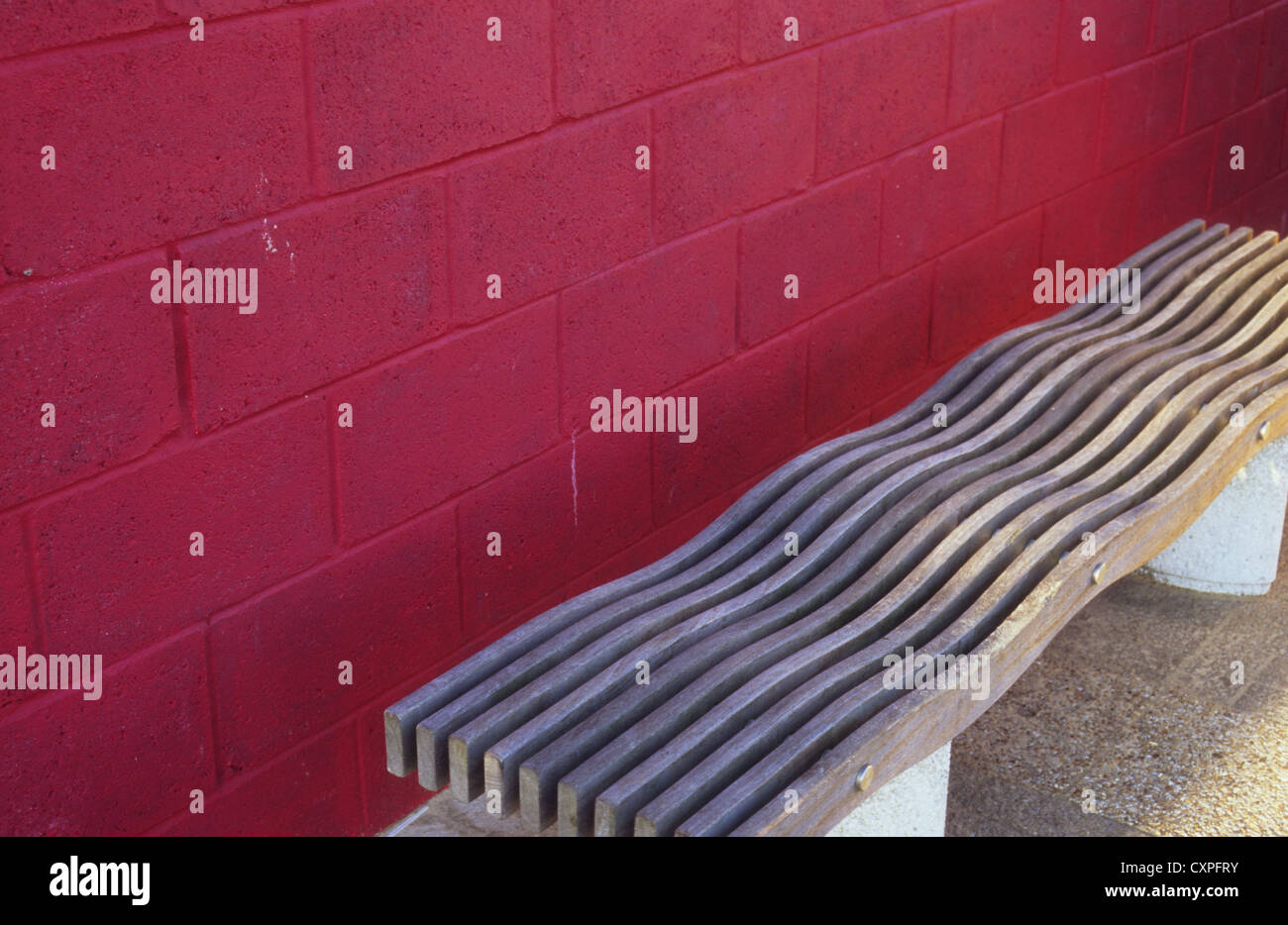 Wooden bench with wavy seat to deter sleepers in front of breeze block wall painted crimson red Stock Photo