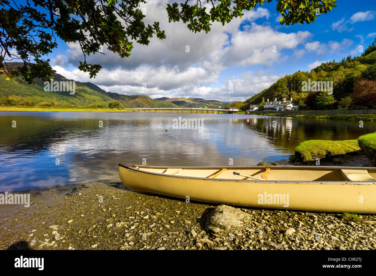 A yellow canoe in the foreground of the river mawddach with reflected blue sky, tree covered hills, white bridge in background Stock Photo