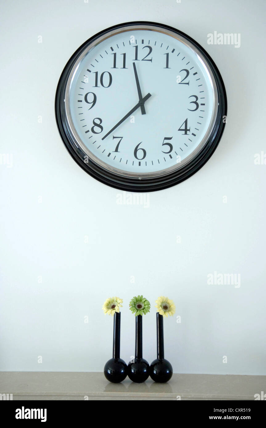 Large traditional wall clock above three small vases with single flowers in each. Stock Photo