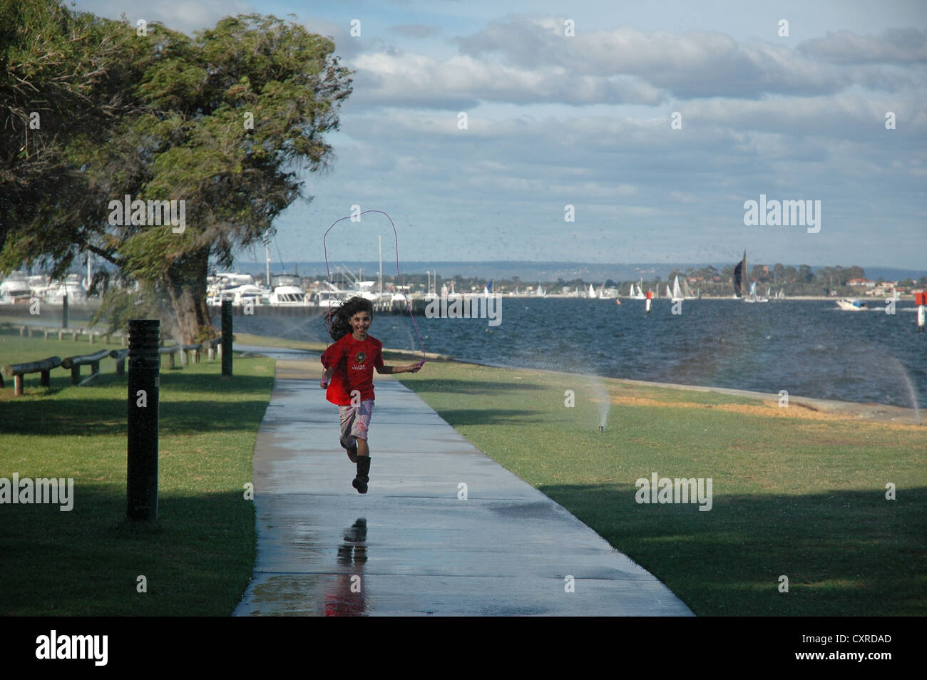 Girl laughing skipping through heavy irrigation shower haze and strong breeze. Swan River, Perth Australia. Stock Photo