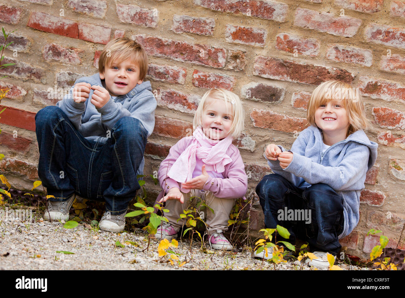 Siblings, 2, 5 and 7 years old, squatting in front of a brick wall Stock Photo