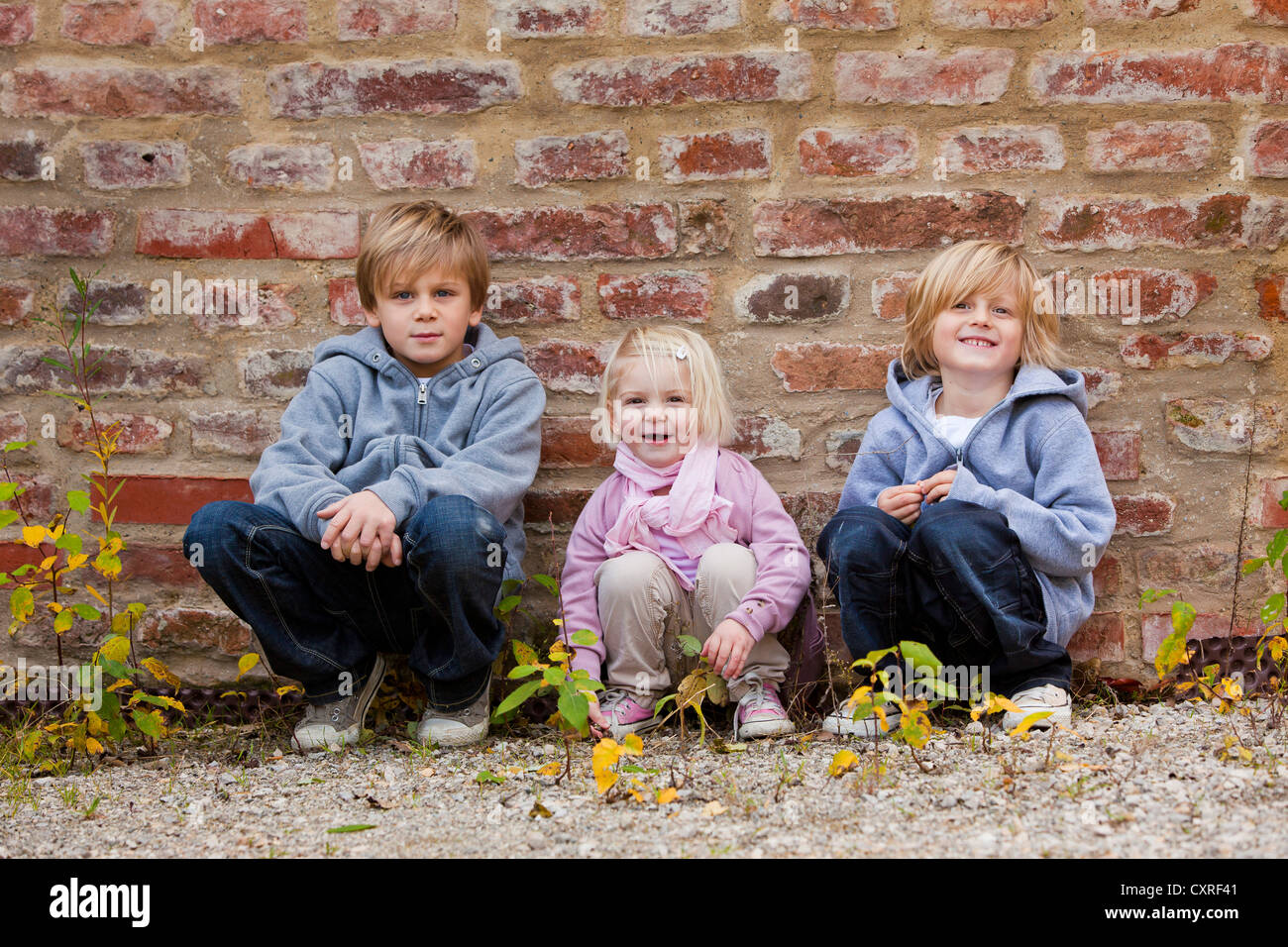 Siblings, 2, 5 and 7 years old, squatting in front of a brick wall Stock Photo