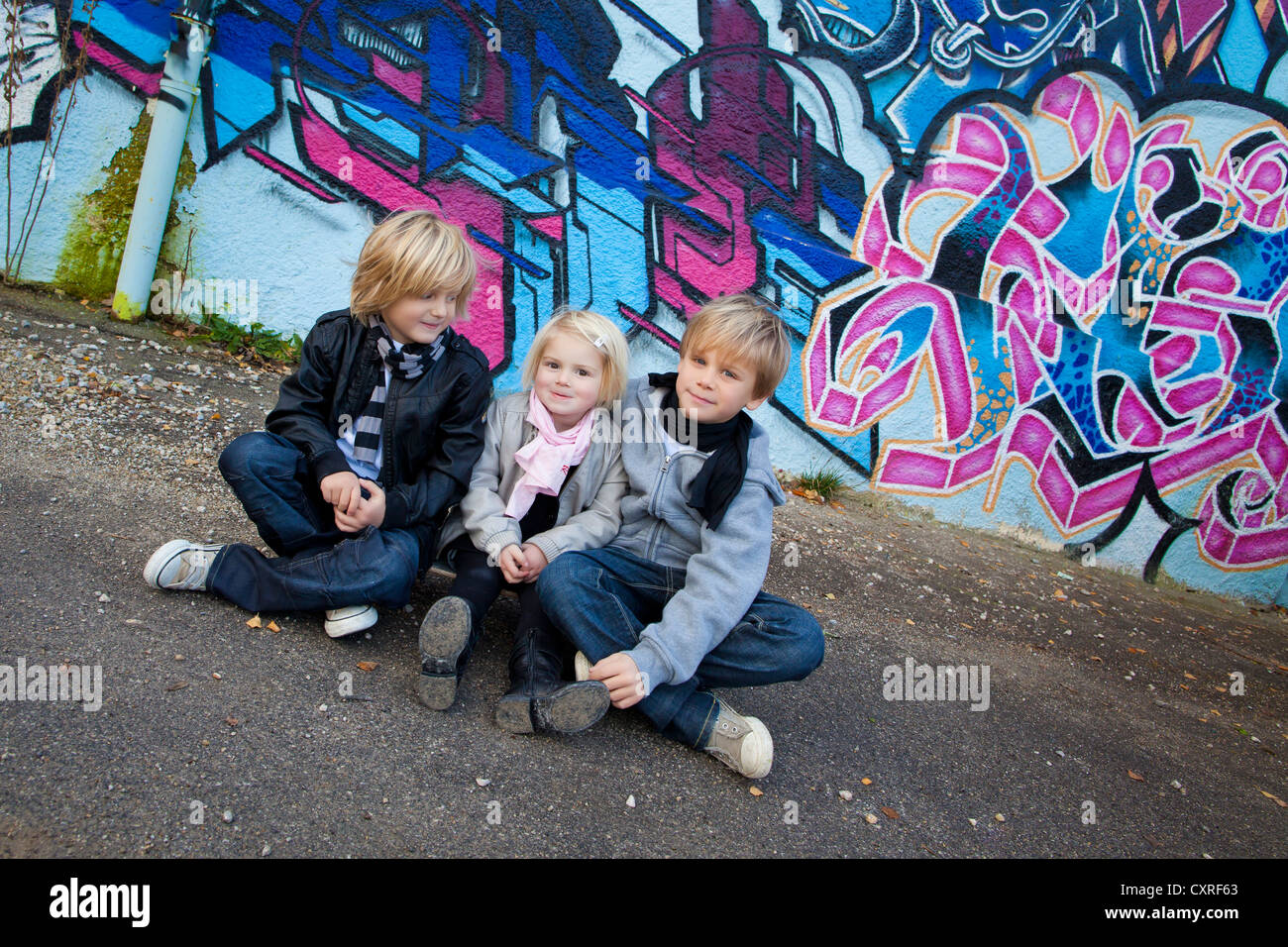Siblings, 2, 5 and 7 years old, sitting on a skateboard in front of a wall with graffiti Stock Photo