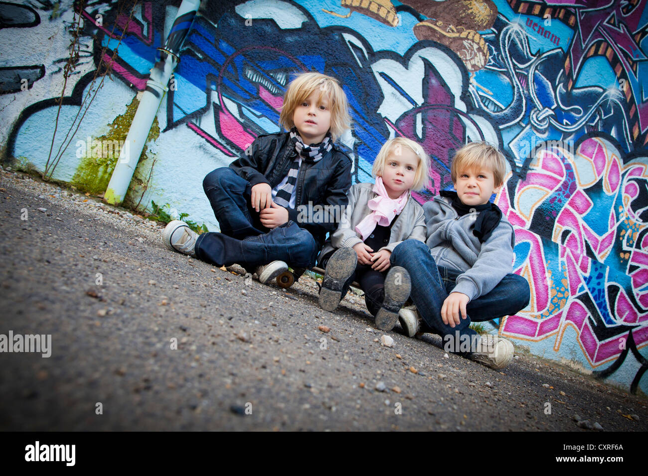 Siblings, 2, 5 and 7 years, sitting on a skateboard in front of a wall with graffiti Stock Photo