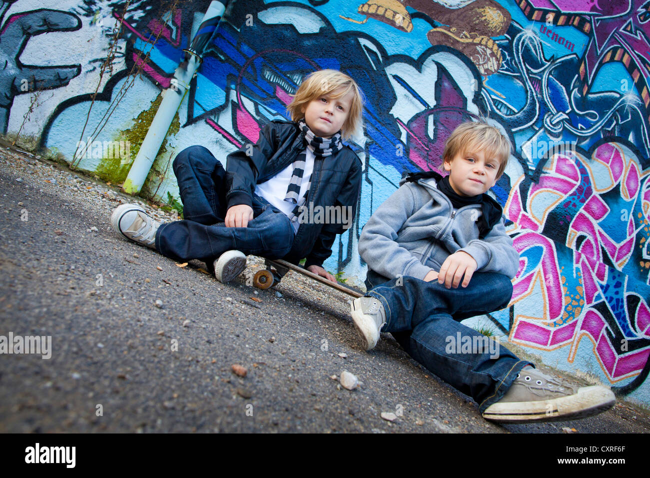 Two boys, 5 and 7 years, sitting on a skateboard in front of a wall with graffiti Stock Photo