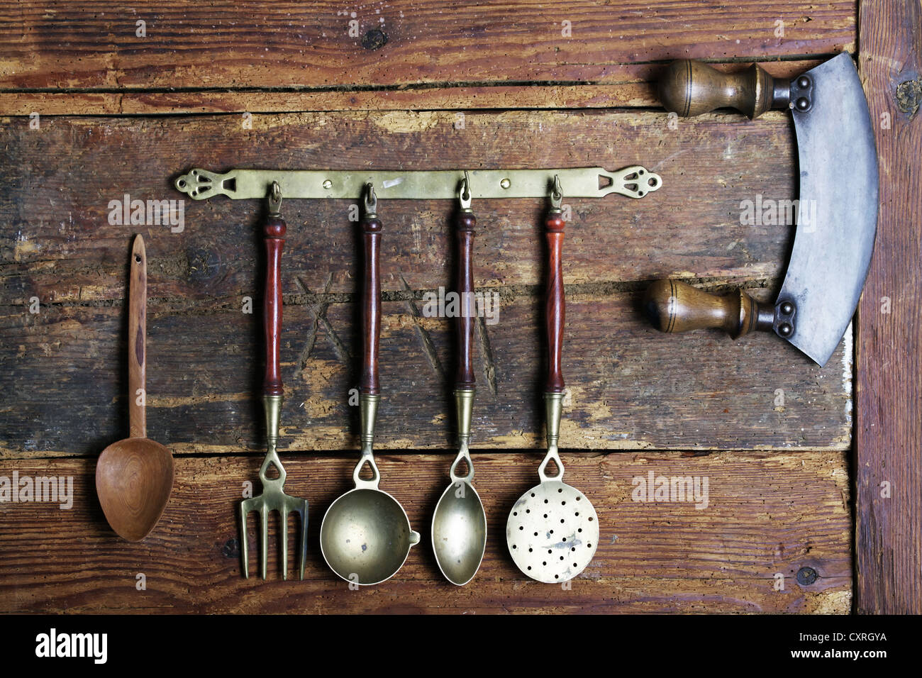 Old kitchen utensils made of brass hanging from a bar in front of a rustic wooden wall Stock Photo