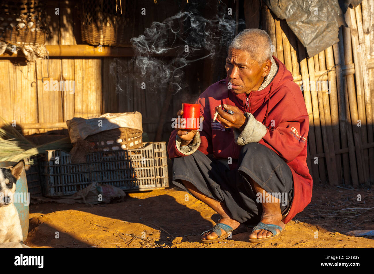 Man smoking a cigarette and drinking tea in front of a straw hut, village of the Akha hill tribe, ethnic minority Stock Photo