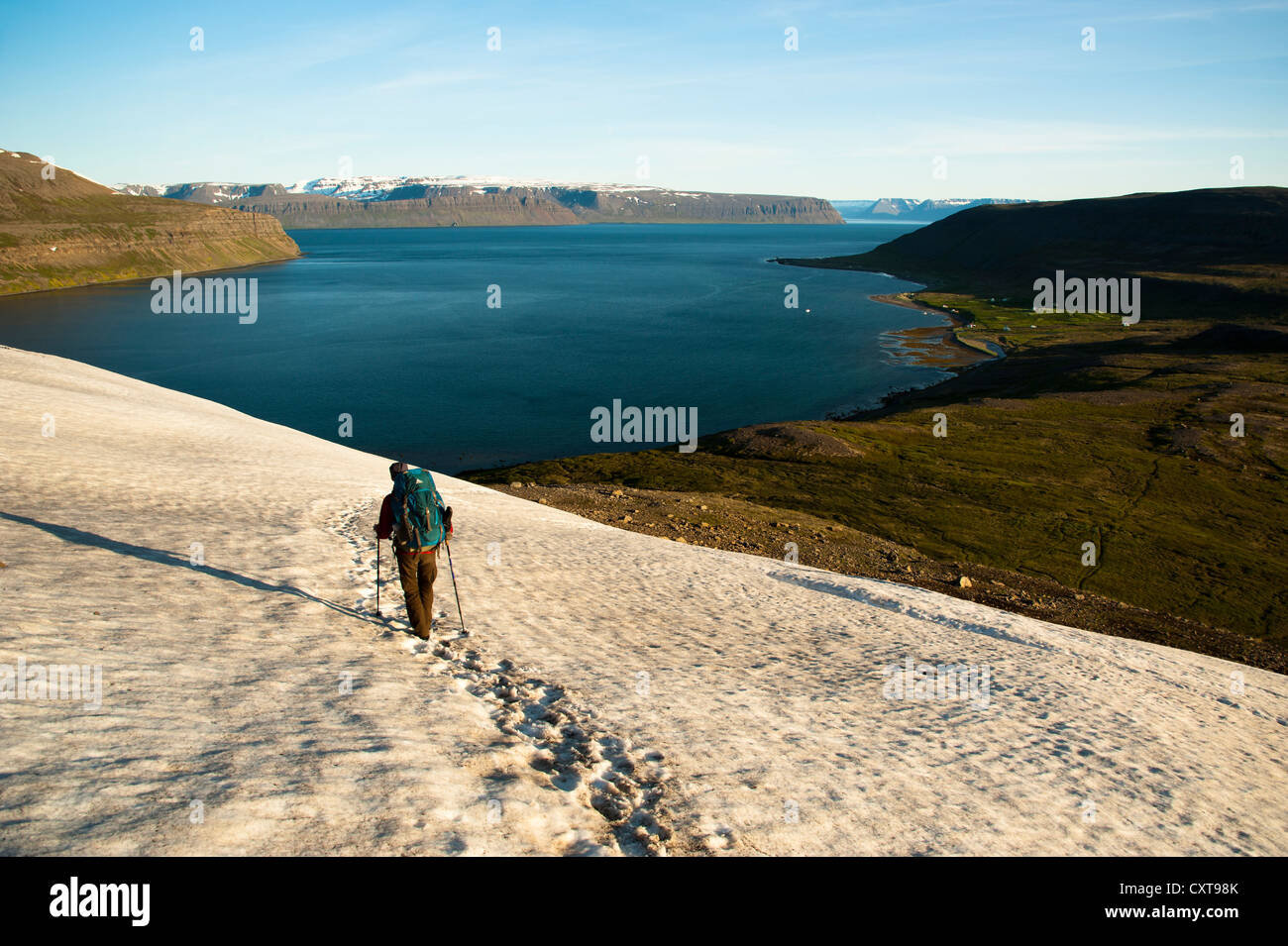 Hiker crossing a snowfield, view of the village of Hesteyri, Hesteyrarfjoerður, Hesteyrarfjoerdur Fjord, Hornstrandir Stock Photo