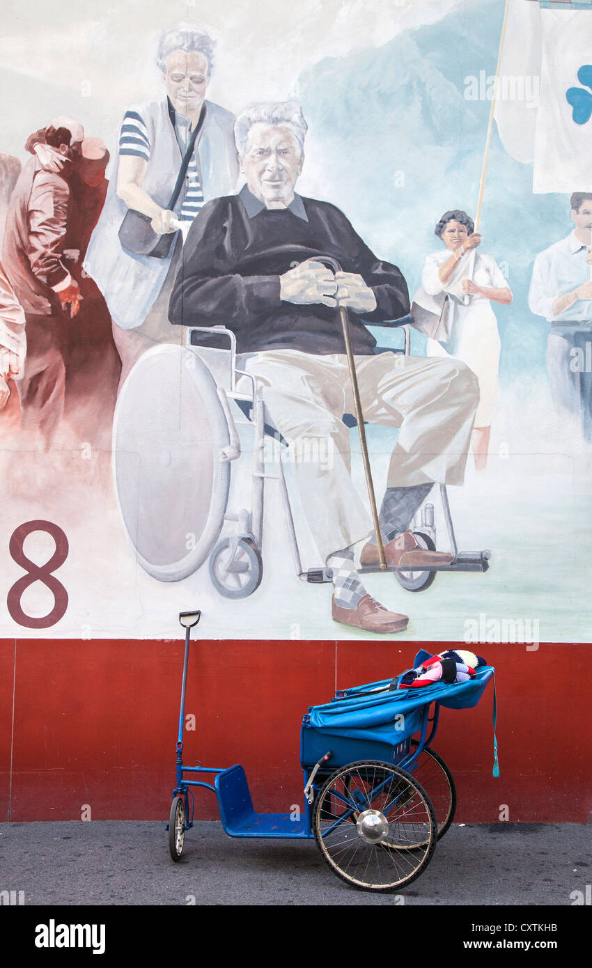 Wheel chair pulled by a Brancadier below a wall painting of a disabled man, Lourdes, Hautes Pyrenees, France Stock Photo