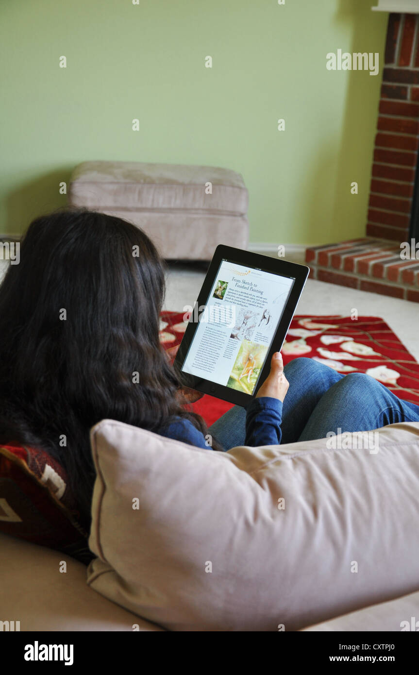 Female student with iPad sitting on sofa at home. An online textbook shown on the iPad screen. Stock Photo