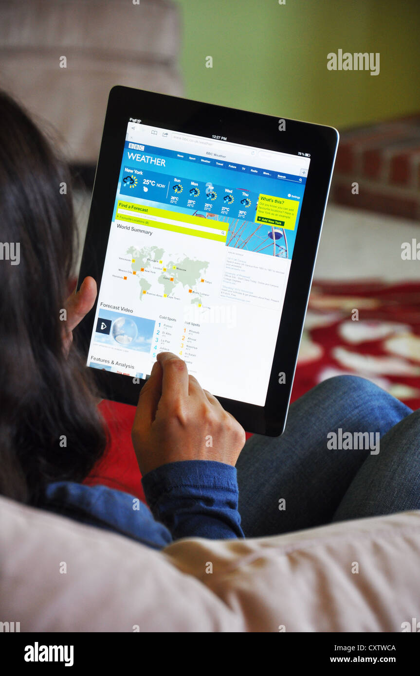 Young girl with iPad sitting on sofa at home. Weather forecast website shown on the iPad screen. Stock Photo