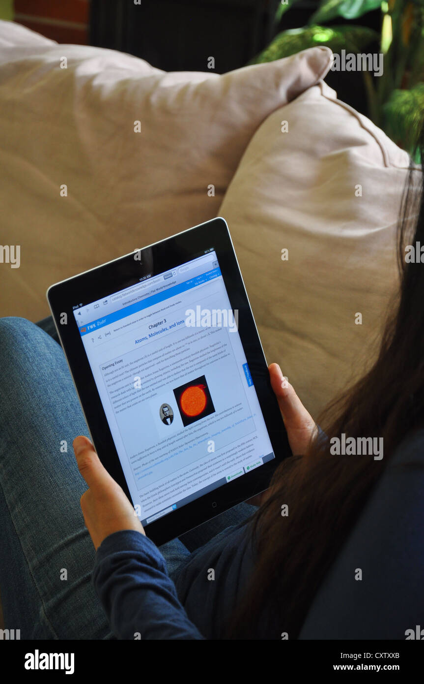 Female student  with iPad sitting on sofa at home. Textbook depicting molecules and atoms shown on the iPad screen. Stock Photo
